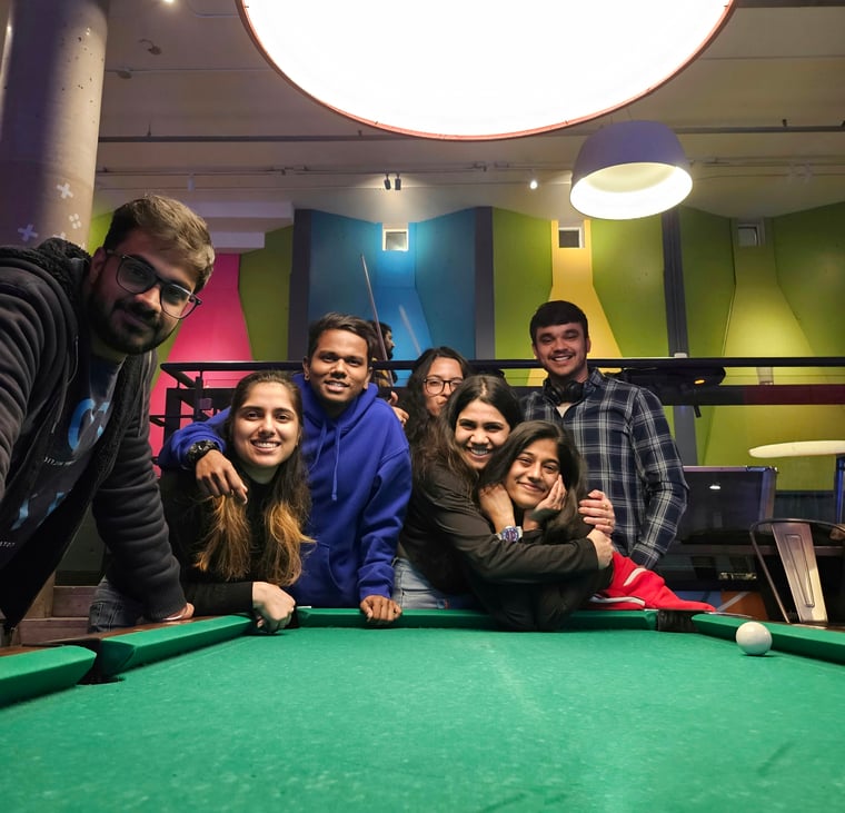 Three boys and four girls standing around a pool table. Two of the girls are hugging each other. All of the individuals are smiling. There are two large bright yellow lamps hanging from the ceiling above the group. The walls in the room are colored in pink, blue , green and yellow. 