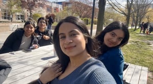 A group of four friends sitting around a table outside on a sunny day. The girl in the front is taking a selfie while smiling at the camera. The three women behind her are smiling too . The background includes the university campus with trees, open sky , and a building.