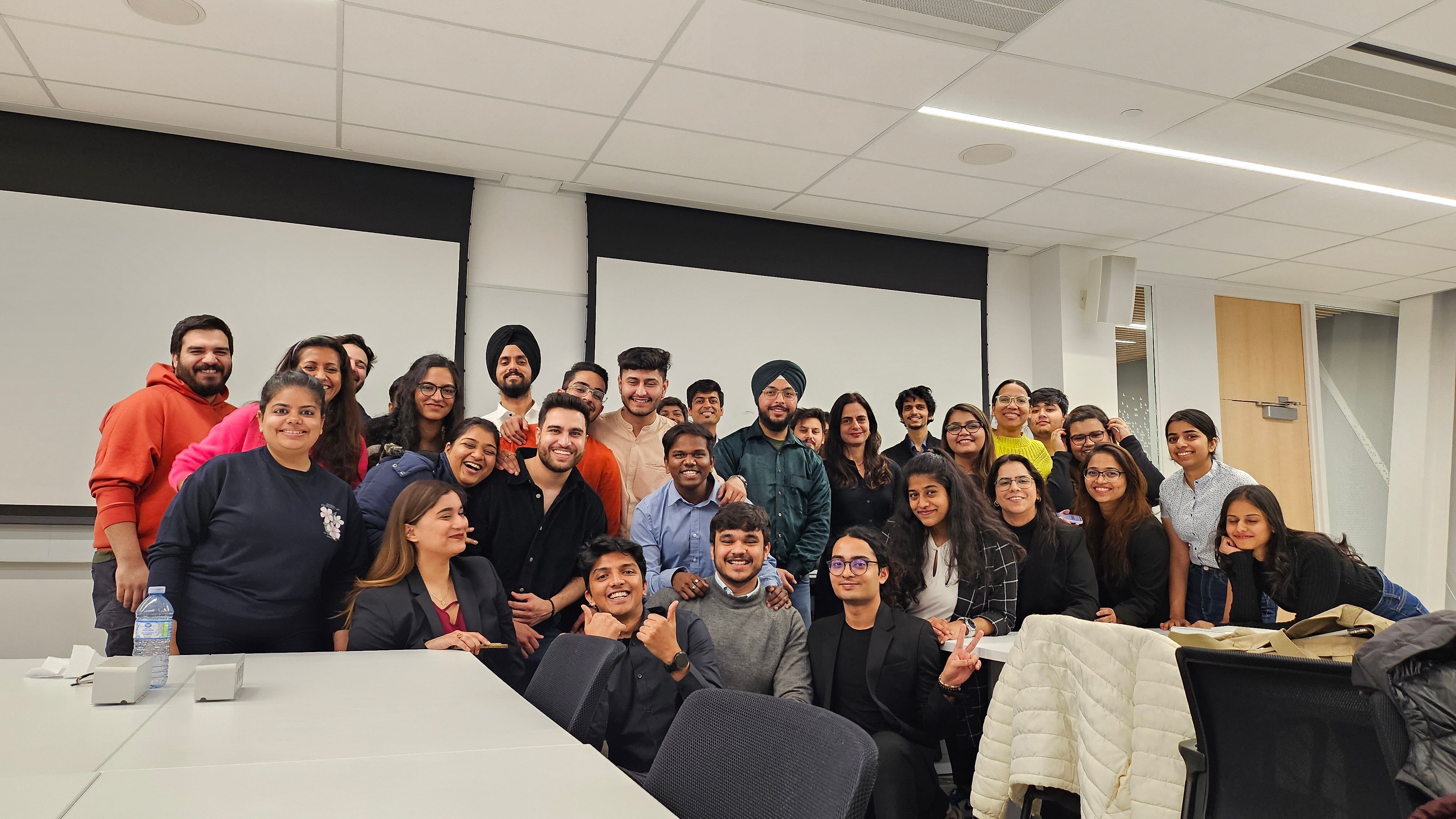 A large group of young people standing together in a group in a classroom. The walls have large whiteboards. The room has long white tables and black chairs. Everyone in the group is smiling
