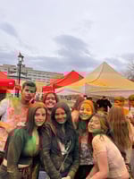 A bunch of people with color on their faces and clothes, with tents and a building in the background during evening time.