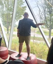 A professor standing on top of the couch, pointing towards the right side of the frame, with trees and bushes in the background behind a glass wall