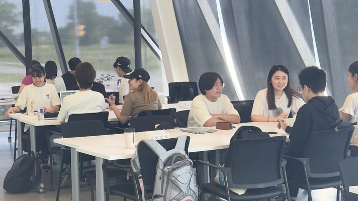 A group of student is seated at tables in a room with large windows and distinctive curved architectural features. The setting appears to be an educational or study environment.