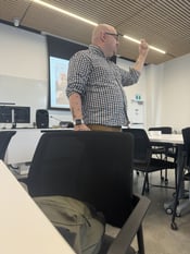 "Professor Tim, a bald and handsome man with tattoos, is seen teaching in a classroom at York University. He is standing and gesturing with his hand, wearing a checkered shirt, while delivering a lecture. The classroom is modern, with chairs and tables arranged for students."