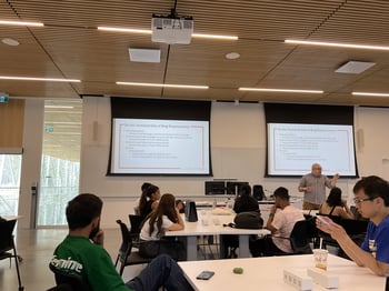 A classroom with students on all the tables, professor standing in the front, and two projector screens with some text on them