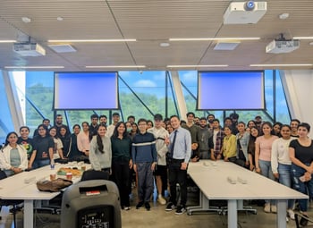A classroom with two projector screens on the back and a bunch of people standing for a group photo behind two desks