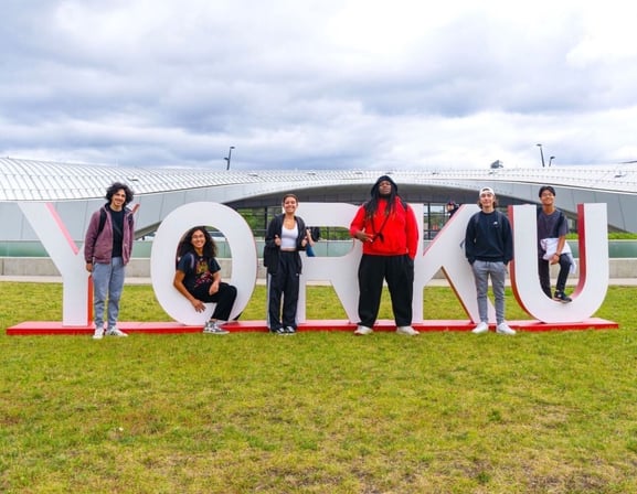 A group of international students posing in front of the York University sign on a green lawn with a modern building in the background.