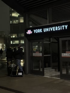 Entrance of the York University subway station at dusk. York University is glowing. There is a man standing beside the entrance playing a trumpet. There is a building behind the station with multiple lit windows. Few students can be seen walking towards that building