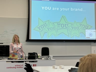 a blonde professor could be seen teaching the class with a big white board behind her. The white board has the slide opened which says You are your brand with 5 green stars and You written inside the stars. 
