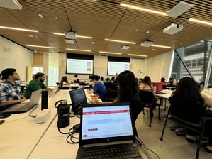 A classroom setting, multiple students are sitting with their laptops on. Few are looking at the presentation on the white board
