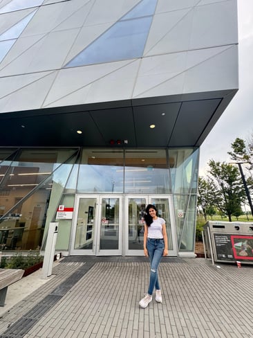 A girl standing in front of a modern building with glass doors, wearing a white t-shirt and jeans, and smiling at the camera.