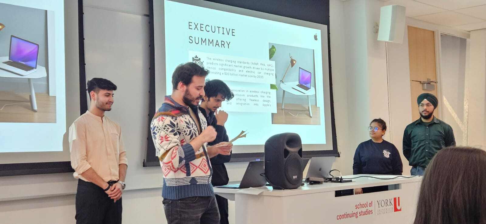 There are 5 people. One of them is holding a paper and looking down towards it. There is a big table York University School of Continuing Studies written on it. There are also one speaker and two laptops and some wire on it. In the background there are 2 projected screens. Executive summary is projected on the screen.