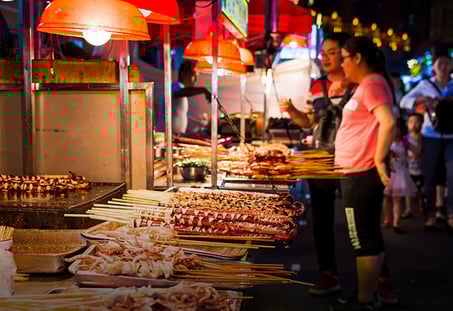 The image shows a vibrant street food market scene at night. There are several skewers of various grilled meats and seafood displayed on trays and grills under red overhead lamps. Two people, one man and one woman, are closely examining the skewers, likely deciding what to buy. In the background, more people and market stalls can be seen, adding to the lively atmosphere.