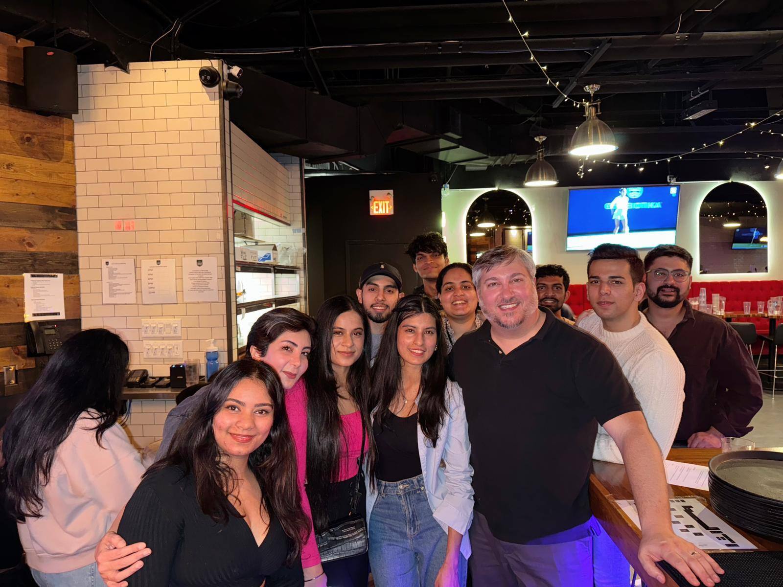 A group of people is posing together inside a restaurant or bar setting. There are ten individuals in the group, including a mix of men and women. They are standing close to each other, smiling and looking at the camera. The restaurant has a modern interior with white tiled walls, wooden panels, and industrial-style lighting. Behind the group, a TV screen is visible, showing a sports event. There is a mix of casual and semi-casual attire, with some people wearing jackets and sweaters. The atmosphere appears friendly and lively.