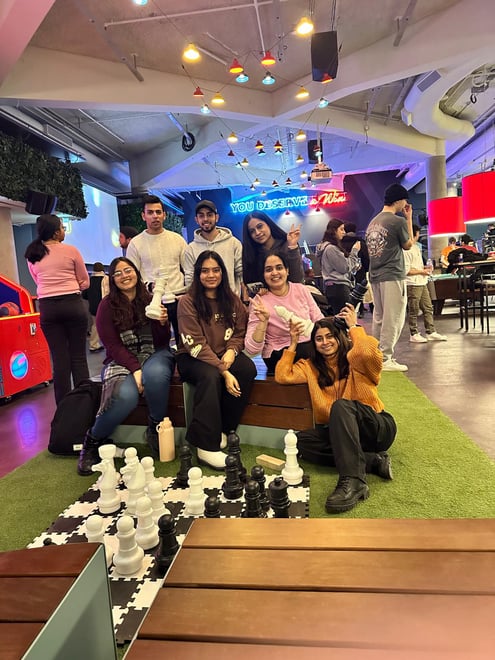 A group of seven people is gathered in an indoor entertainment venue, possibly an arcade or recreation center. They are sitting and standing around a large floor chessboard with oversized chess pieces. The group consists of four women sitting or crouching in the front and three men standing behind them. Everyone is smiling and appears to be enjoying their time together. The venue has a modern, vibrant atmosphere with colorful lighting, hanging pendant lights, and a neon sign in the background that reads, "YOU DESERVE A WIN." Other people can be seen in the background, engaging in various activities. The setting includes a mix of casual and comfortable seating with benches and a green turf floor. Some individuals are holding drinks, and the overall mood is lively and fun.