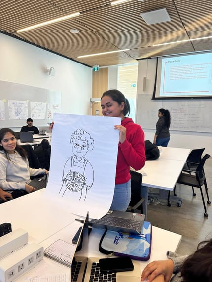In a modern classroom with a wooden ceiling and bright lighting, a young woman in a red hoodie and jeans is standing and holding up a large sheet of paper with a hand-drawn illustration. The drawing depicts a character with curly hair and glasses, holding a pizza with several slices visible. Seated at the table in front of her, another woman in a light sweater looks on, smiling. The table is cluttered with laptops, notebooks, and various other items, suggesting an active and engaging class activity. In the background, other students are working at their desks, and a large screen displays a presentation titled "Pizza Game." A whiteboard with notes and drawings is also visible behind the students. The atmosphere is collaborative and educational.