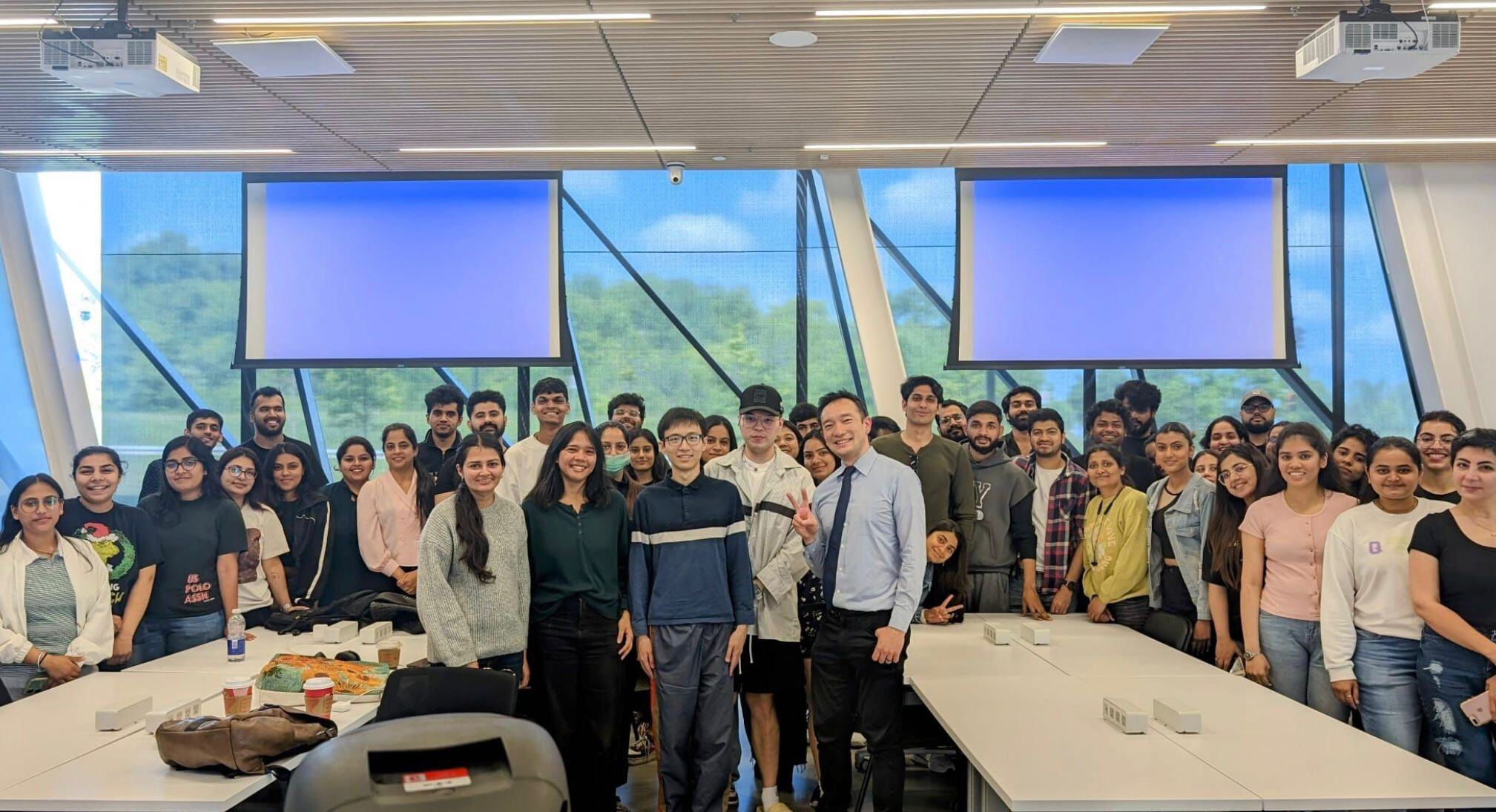 A group photo of students with a guest lecturer at York School of Continuing Education. The group is gathered in a modern classroom with large windows, natural light, and two projection screens.