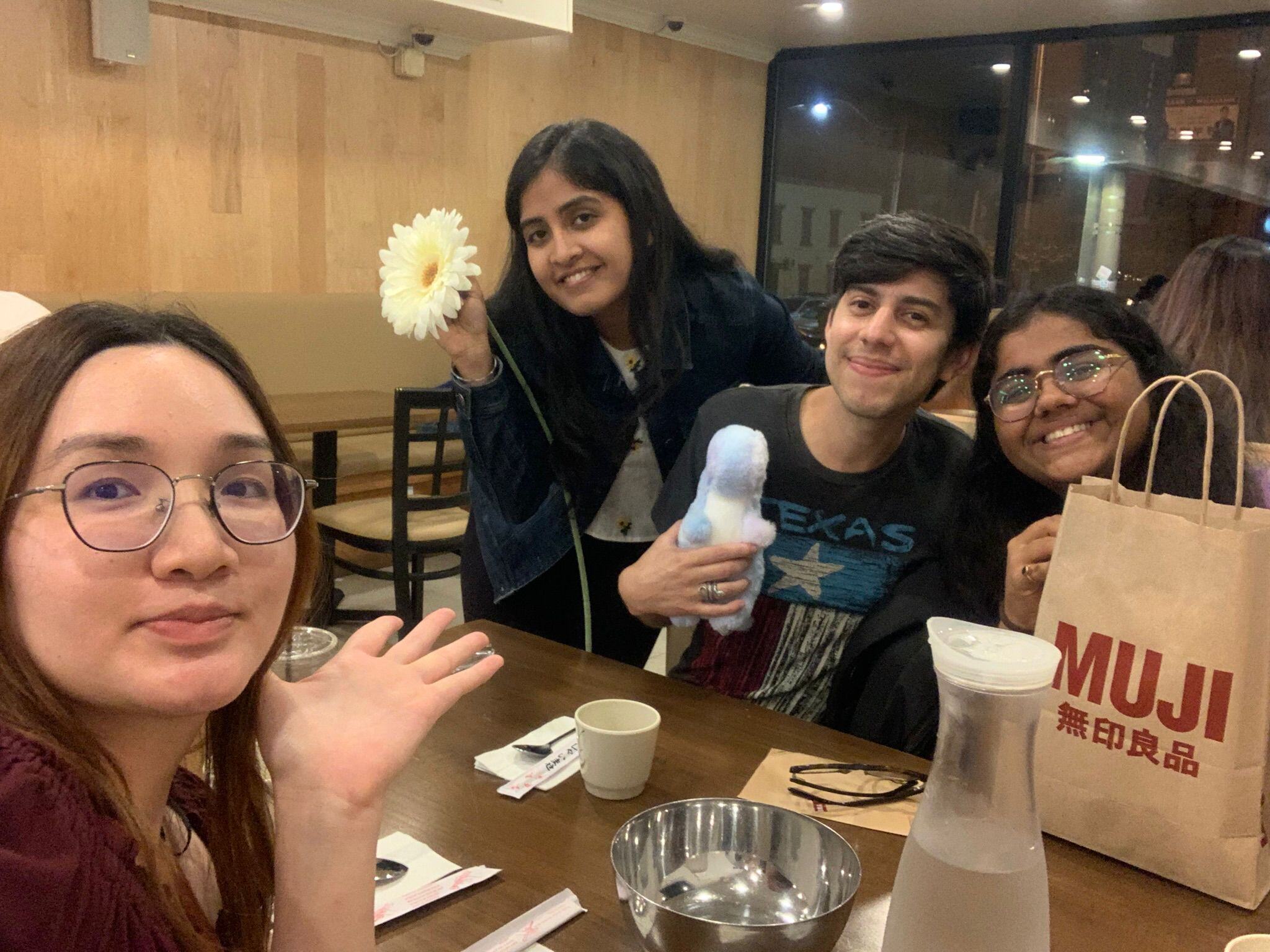 Me with my friends enjoying a cozy dinner together at a restaurant. We are smiling and relaxed, I'm holding a flower and my one friend is holding a plush toy, while a shopping bag from MUJI sits on the table.