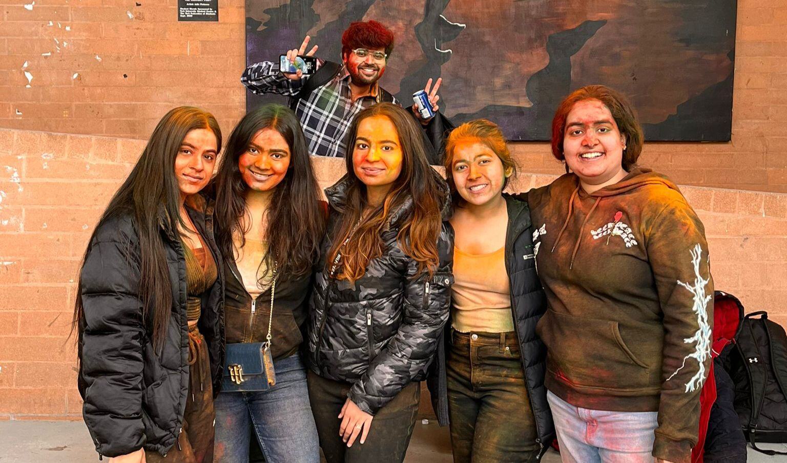 Five young women playing Indian Festival Holi at an international university in Canada, York University. The young adult girls are wearing black clothes and looking straight in the camera with all their faces covered in different colors of the Holi festival.