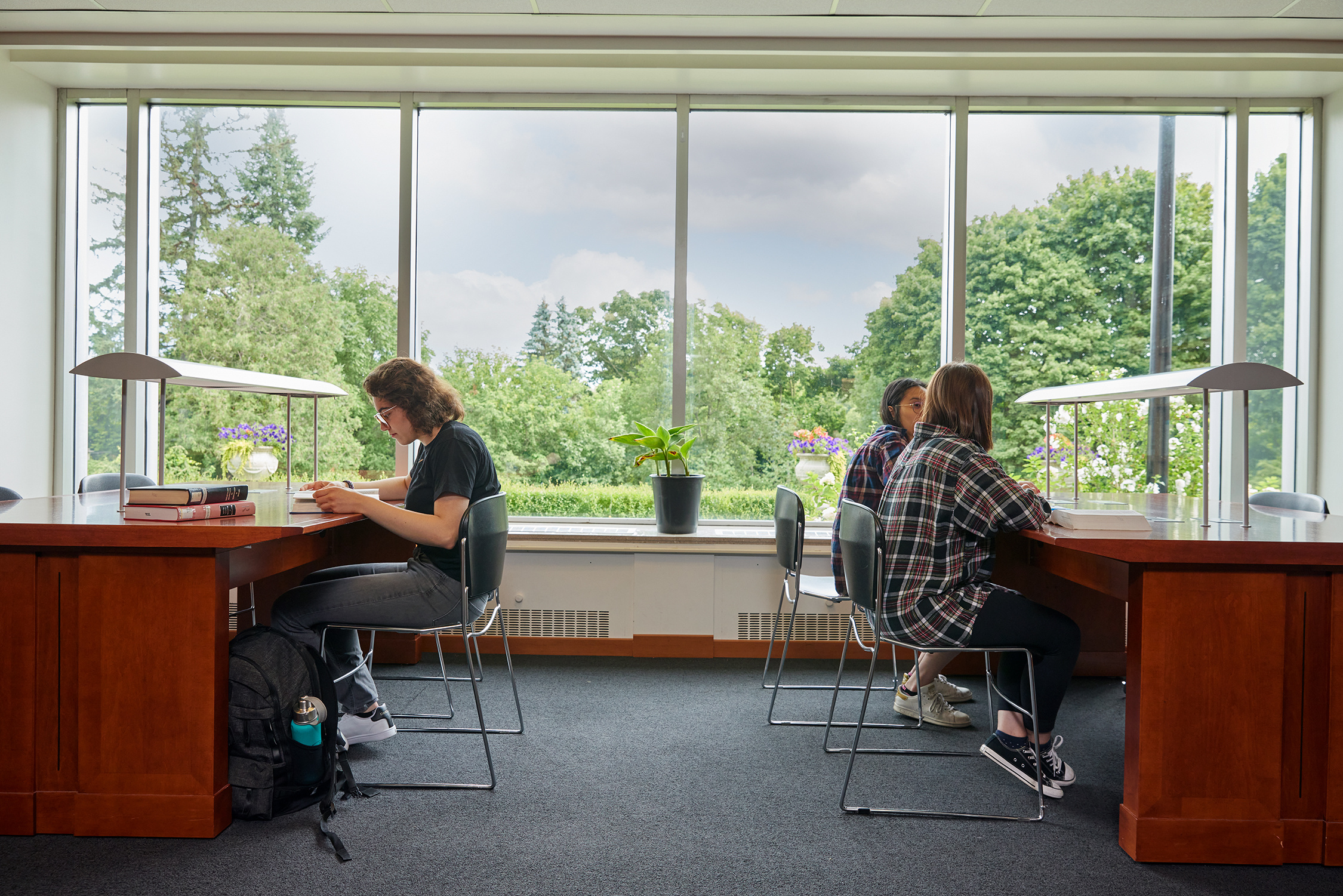 Inside the York SCS building, a student on the left is sitting and reading a book. On the right desk, two students are talking to each other. There's a window at the center showing green trees and blue skies in the background 