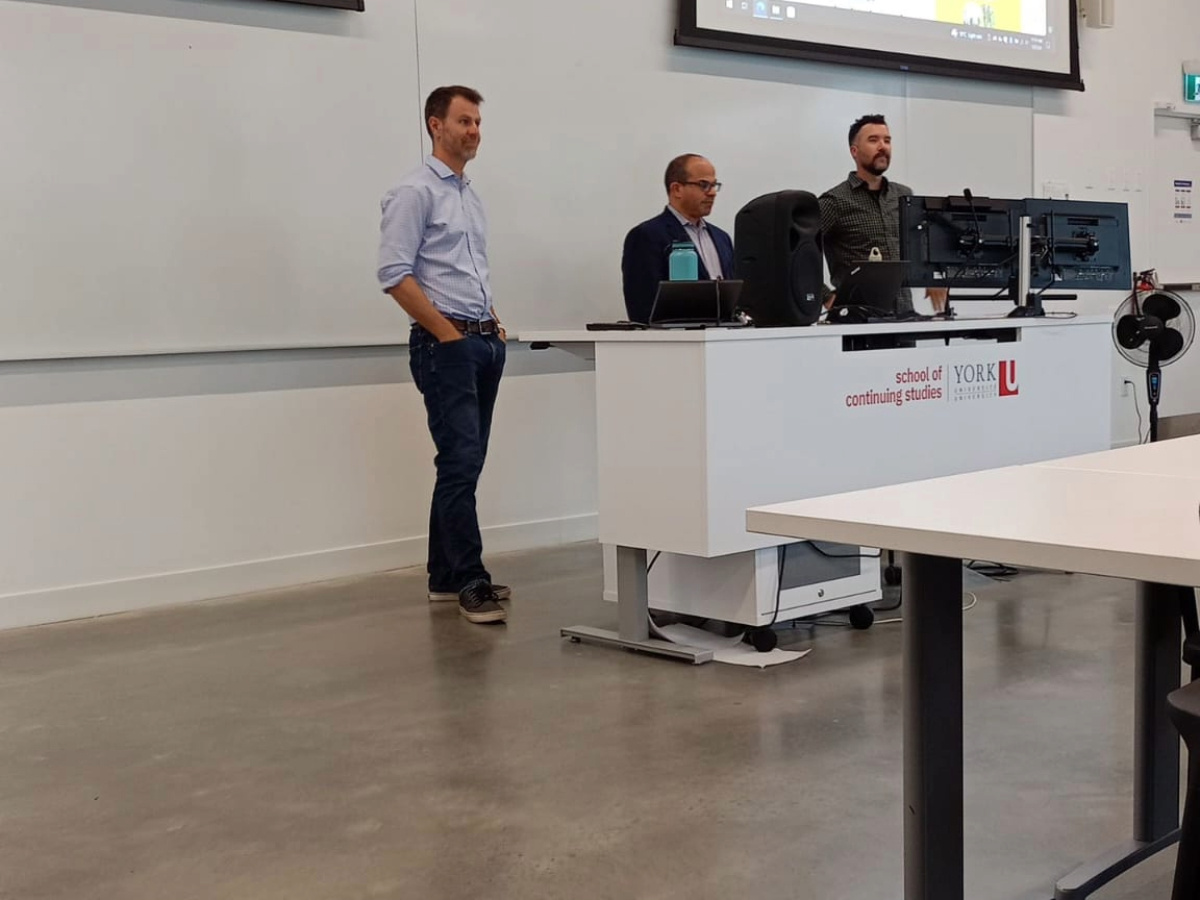 Professor Andrew Temes standing behind the table with two guest lecturers. There are computers attached on the table. Upper side of the frame there something being shown through a projector.