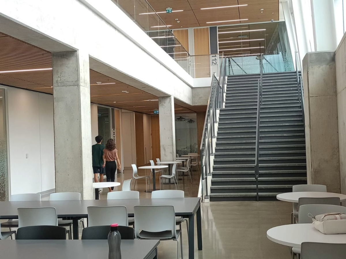 York University school of continuing studies inside campus showing ground floor. A cafeteria in the frame with lots of tables and chairs. On one of the tables there is a bottle, on the right side of the frame there stairs that leads up to first floor.