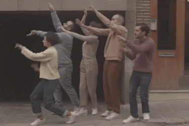 Group of 5 people waving their hands on a sidewalk in front of a building