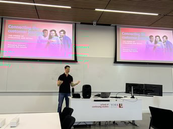 Uri Gorodzinsky, Managing Director of Amazon Ads Canada, delivering a guest lecture on 'Connecting the Customer Journey: The Power of Streaming and Retail' at York University School of Continuing Studies. The presentation slide is displayed on two large screens, and Uri is standing at the front of the classroom adressing the students