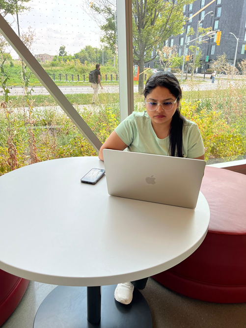I am  working on a laptop at a round table in a bright, modern study space with large windows and a view of the outdoors at York University