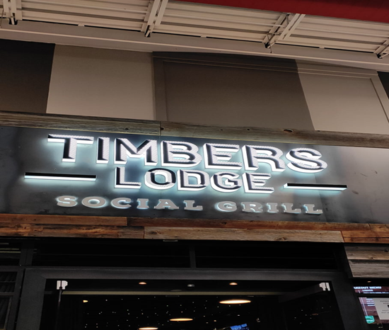 Entrance sign for Timbers Lodge Social Grill, featuring illuminated lettering against a rustic wooden backdrop