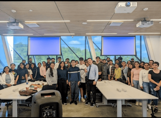 A group of students stands together for a group picture in a room with large windows that let in ample natural light. The room is equipped with projectors mounted on the ceiling, and there is a square table in front of the group. The students are smiling and appear to be part of a class or workshop.