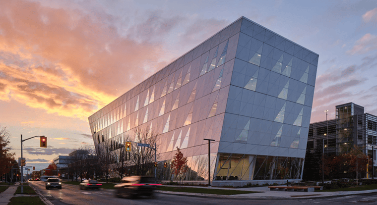 A big building with large glass triangular windows . There are trees by the road with little to no leaves. Cars are speeding away on the road. 