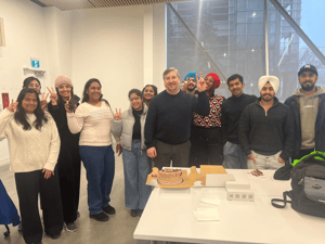 "Professor Heath, standing in the center, is surrounded by a group of students in a classroom at York University, celebrating his birthday with a cake on the table in front of them. The group is smiling and making peace signs, creating a cheerful and festive atmosphere. The classroom has large windows, allowing natural light to fill the space."