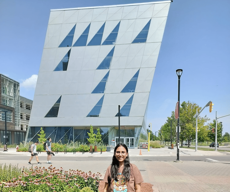 “The author stands smiling in front of the York University school of continuing studies building. Its a sunny day with blue sky, flowers, trees, cars and few pedestrians in the background.”