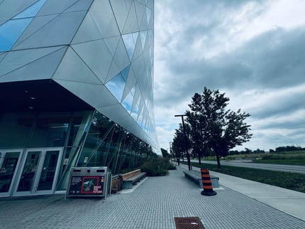 Photo of the Continuing Studies building at York University. The left half shows a modern building, while the right half features nature with trees and a long road. A trash can and two benches are on the left side.