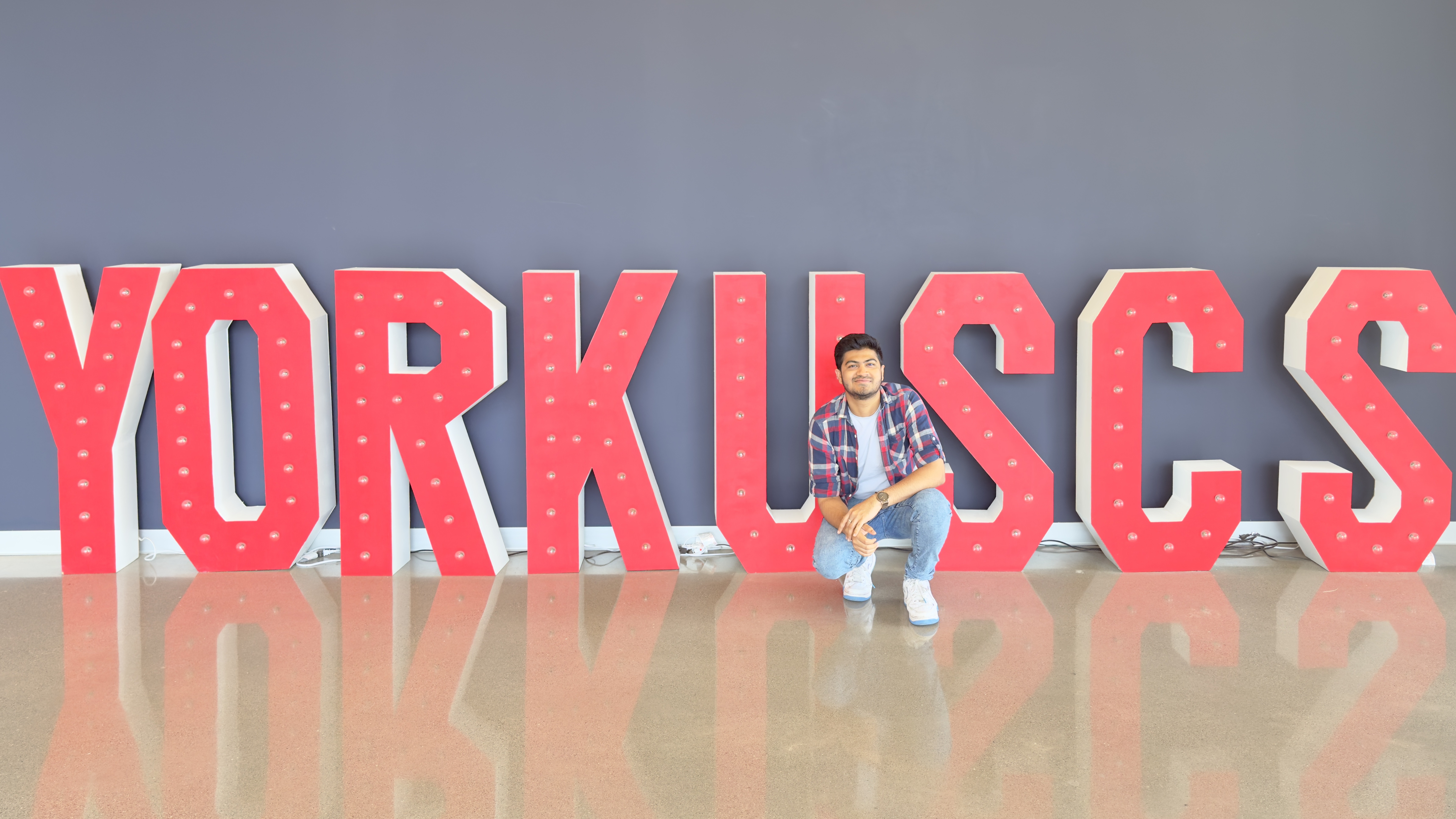 A person is sitting on the floor in front of large red letters that spell out “YORKUSCS” indicating an indoor setting possibly related to York University’s School of Continuing Studies. The background is a plain grey wall 
