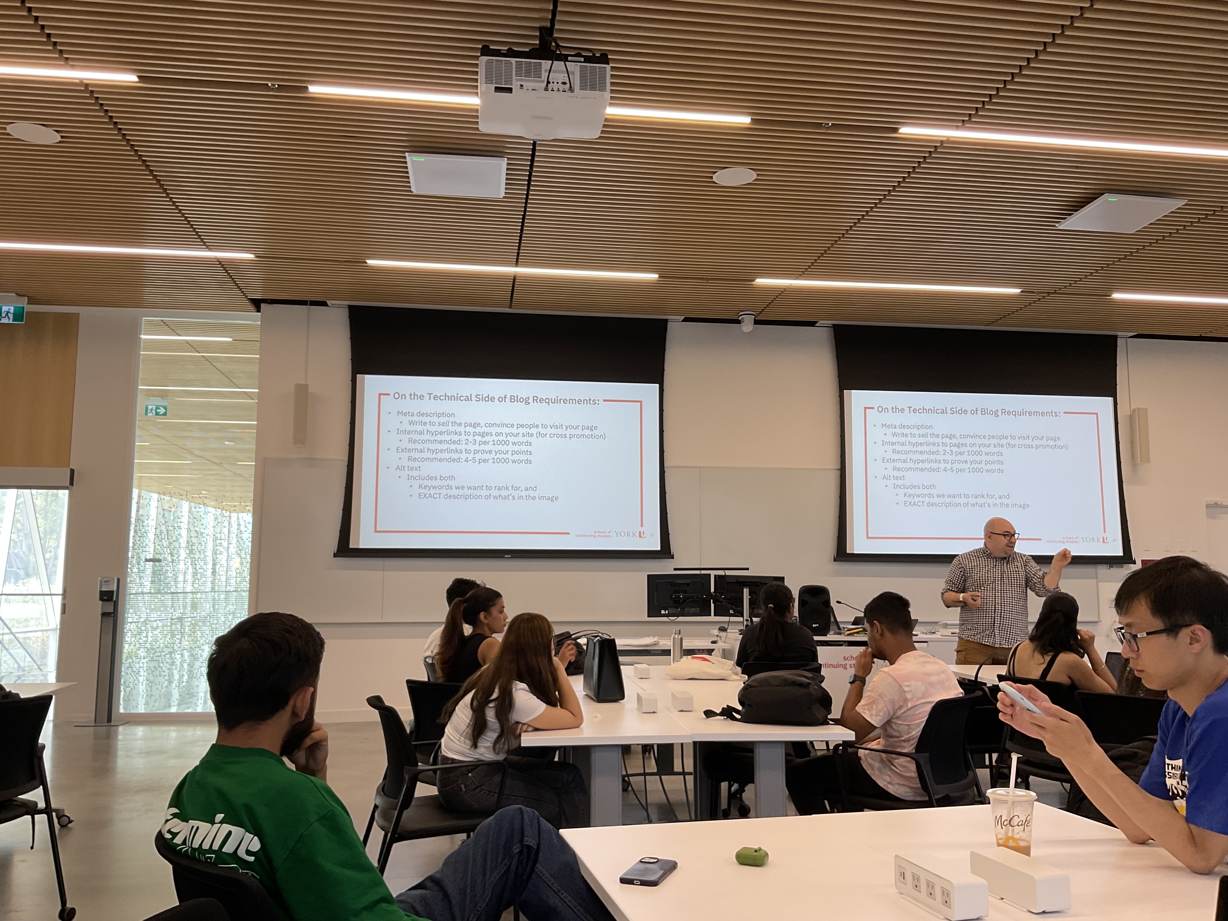 A classroom in the school of continuing studies building, with students sitting around the tables, and a professor in front of the class with two projector screens in the background