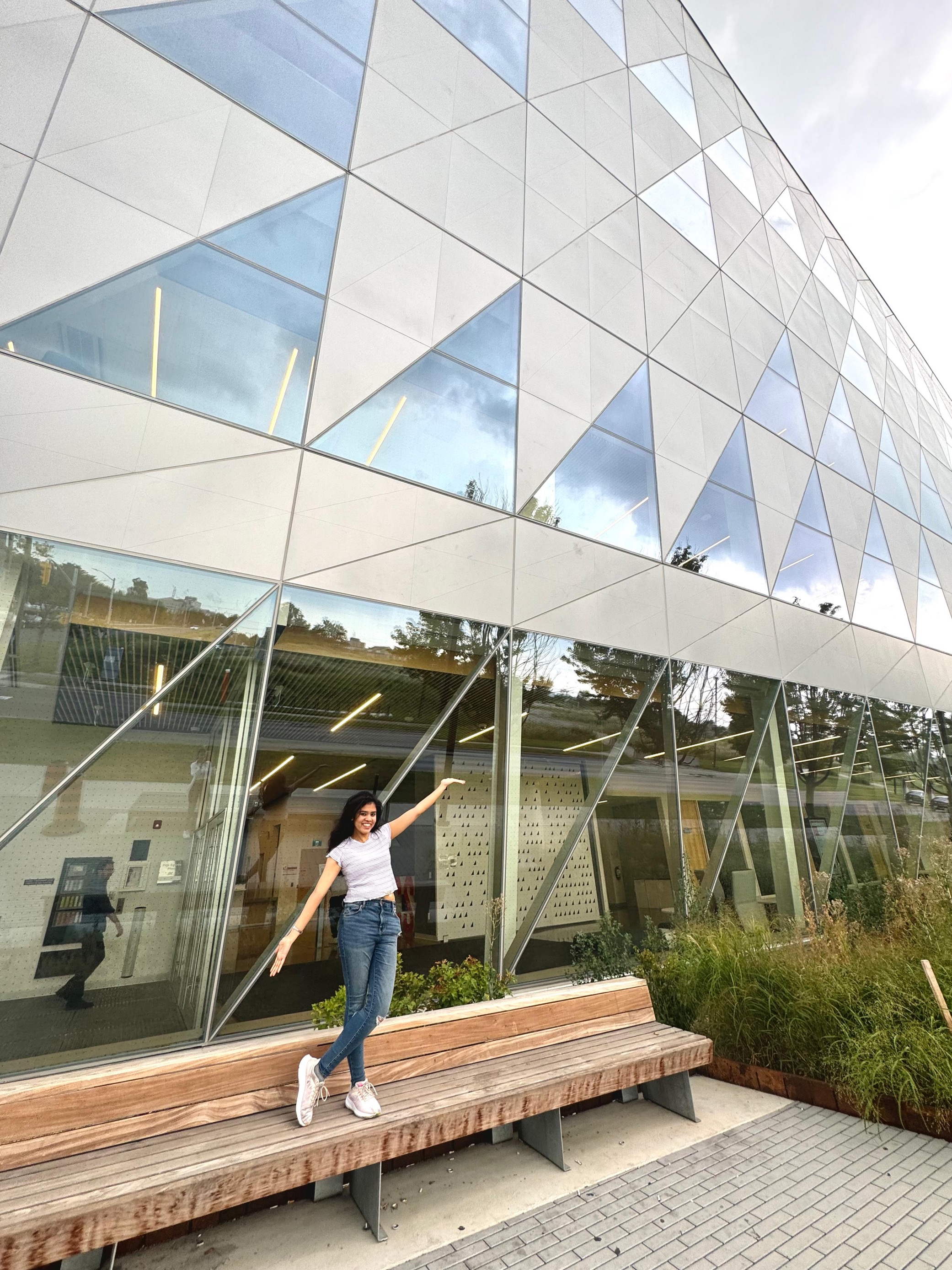 A girl standing and posing cheerfully in front of a glass building with geometric patterns, wearing a white t-shirt and jeans.