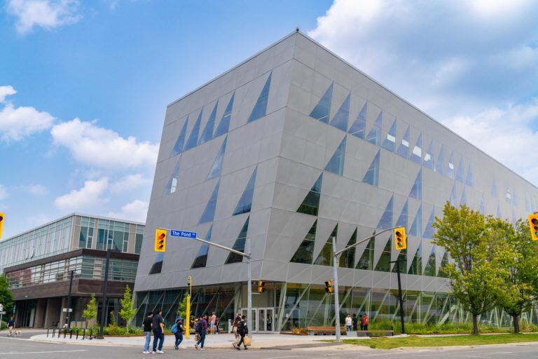 The image portrays York University School Of Continuing studies,modern, geometrically designed building with glass panels under a clear blue sky. The urban scene is animated with pedestrians crossing the street in front of this architectural marvel.