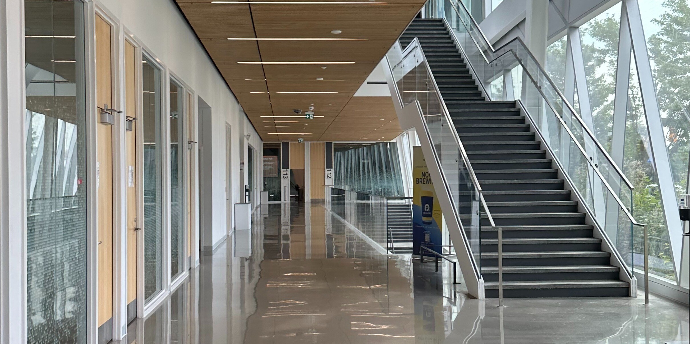 A spacious, well-lit interior corridor of York University's School of Continuing Studies. The hallway has a wooden ceiling with linear light fixtures, glass walls on one side, and a staircase leading to the upper floor.