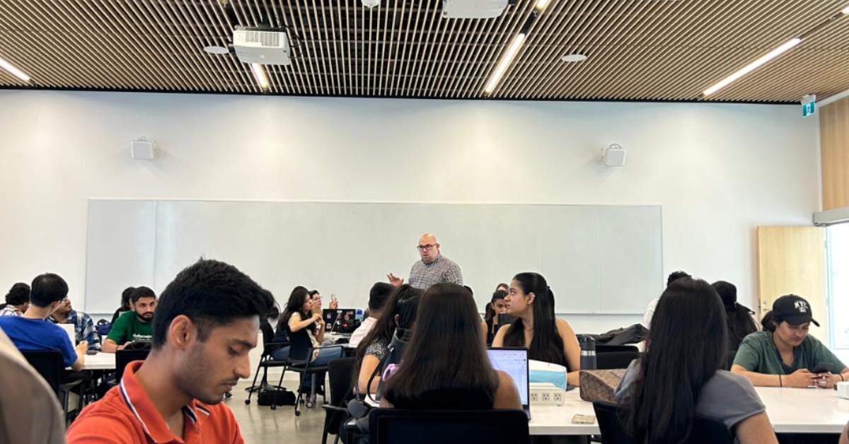 Classroom at York School of Continuing Education with students seated at tables, a professor standing and speaking at the front, and a ceiling with wooden slats and modern lighting fixtures